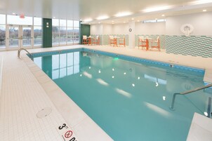 Piscina interior con suelo de baldosas, una escalera de piscina y un letrero que indica prohibido bucear. Hay sillas y mesas a lo largo de la piscina.
