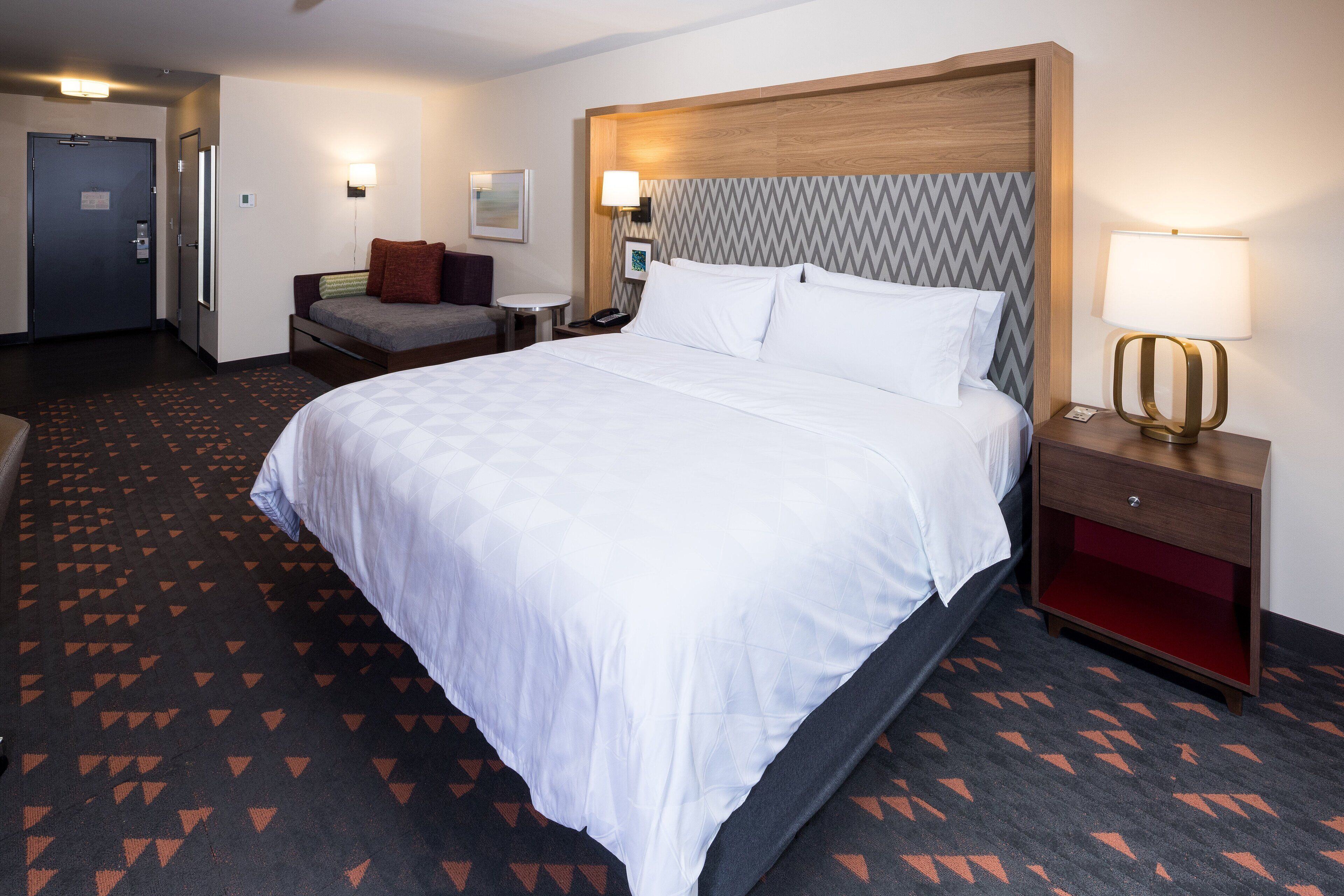 A hotel room with a large bed, a bedside table with a lamp, a small round table, and a chair.