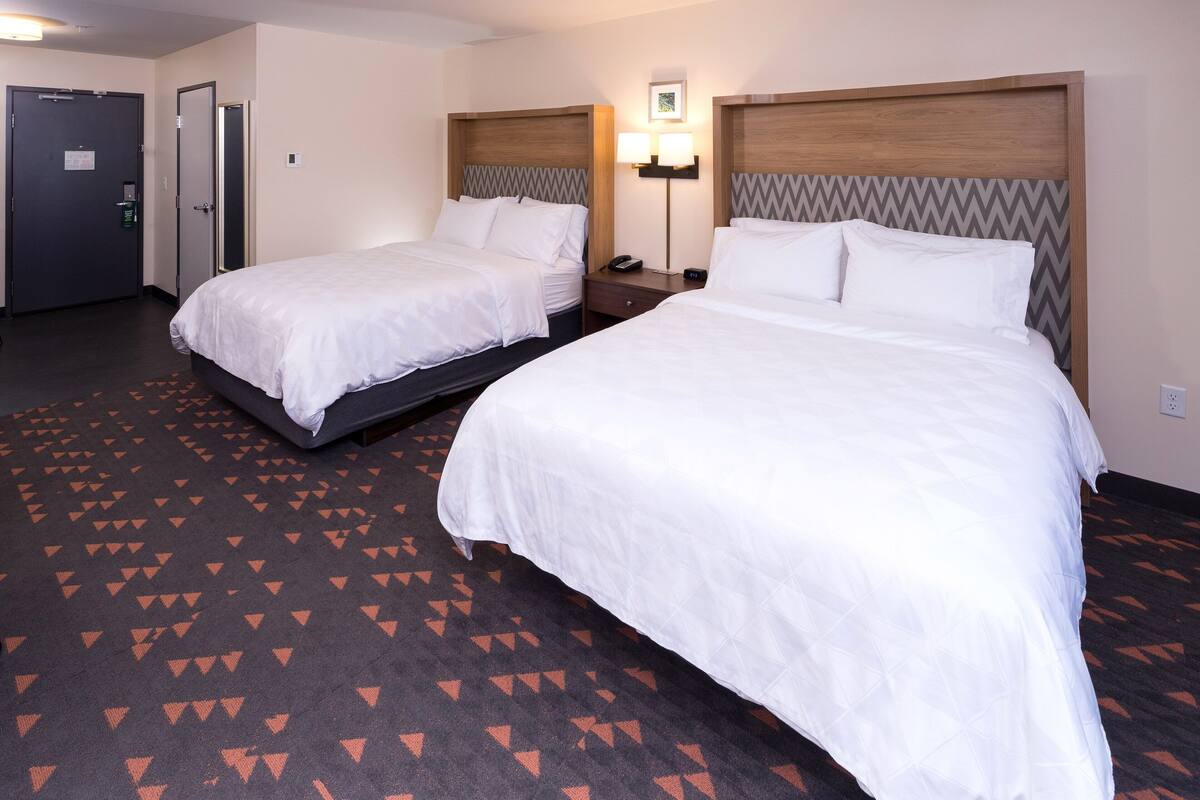 A hotel room with two beds, a carpet with a geometric pattern, a wall-mounted lamp, and a door with a security lock.