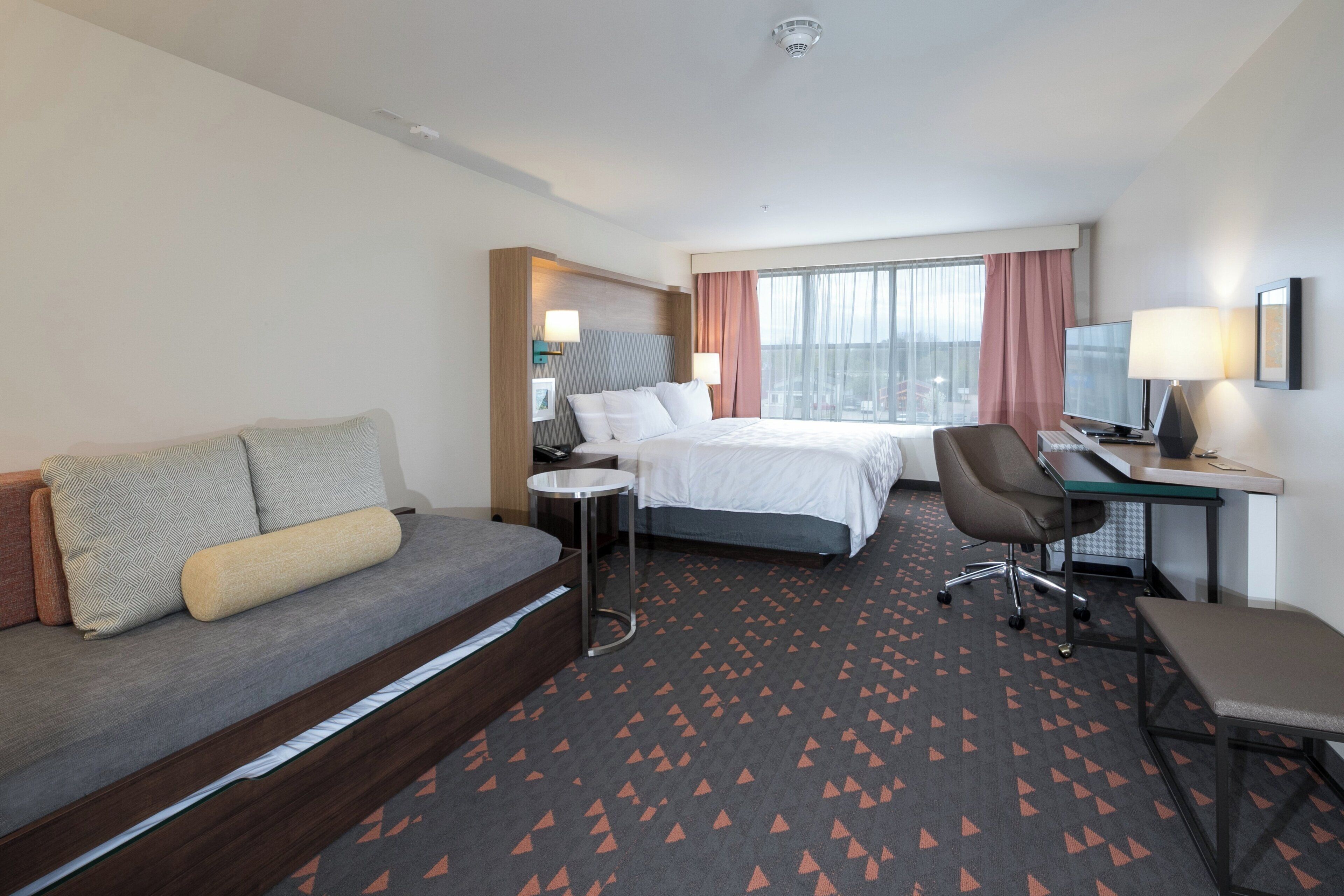 Una habitación de hotel con cama, escritorio, silla, sofá y ventana con cortinas.