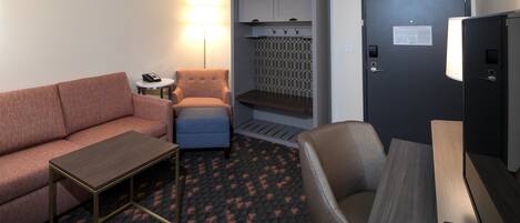A hotel room with a sofa, armchair, small table, desk, and a cabinet with hooks.