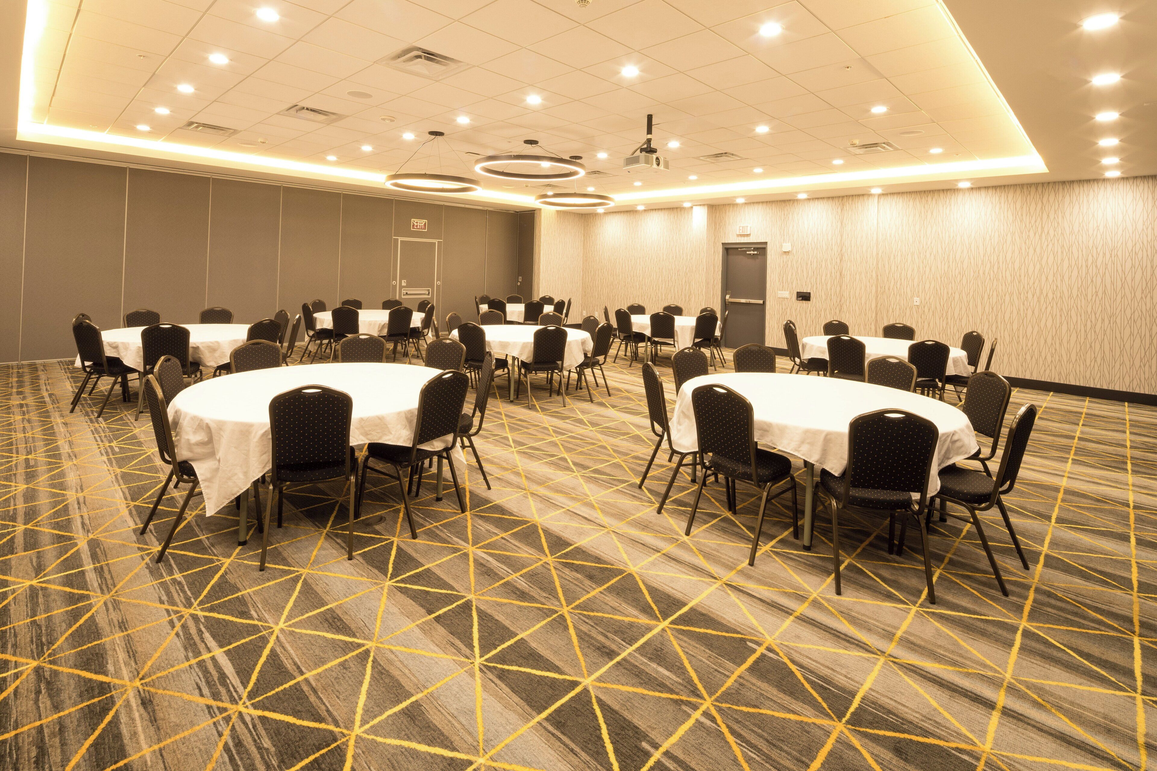 Une salle de conférence avec des tables et des chaises rondes, un sol recouvert de moquette et un projecteur fixé au plafond.