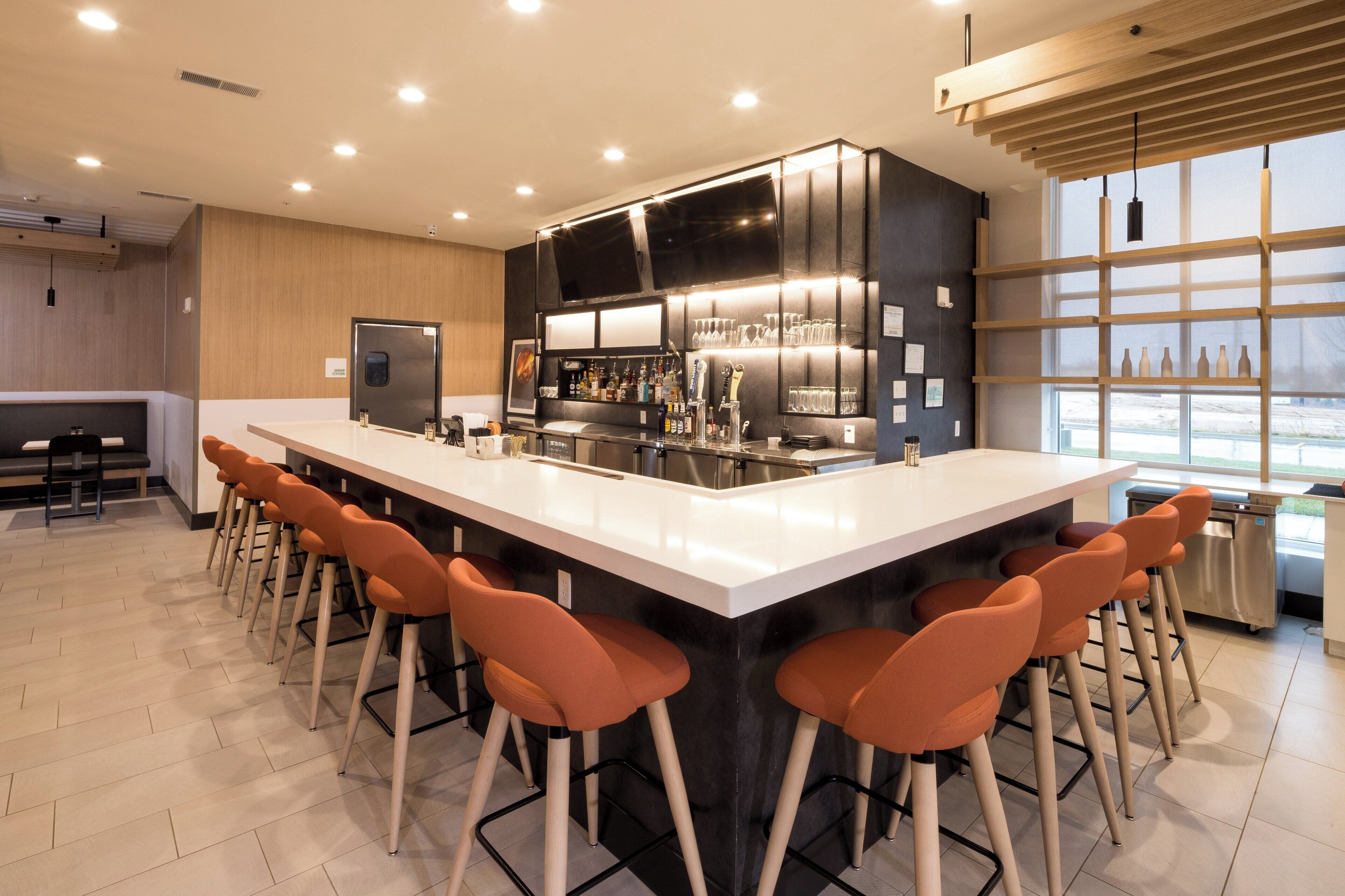 Zona de bar moderna con una barra larga, taburetes y un área de cocina con refrigerador y equipamiento de bar.