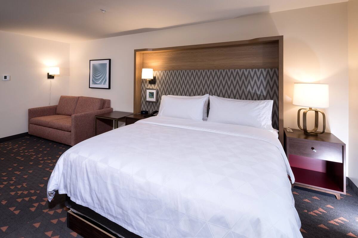 A hotel room with a large bed, a brown armchair, a nightstand with a lamp, and a wall-mounted picture.
