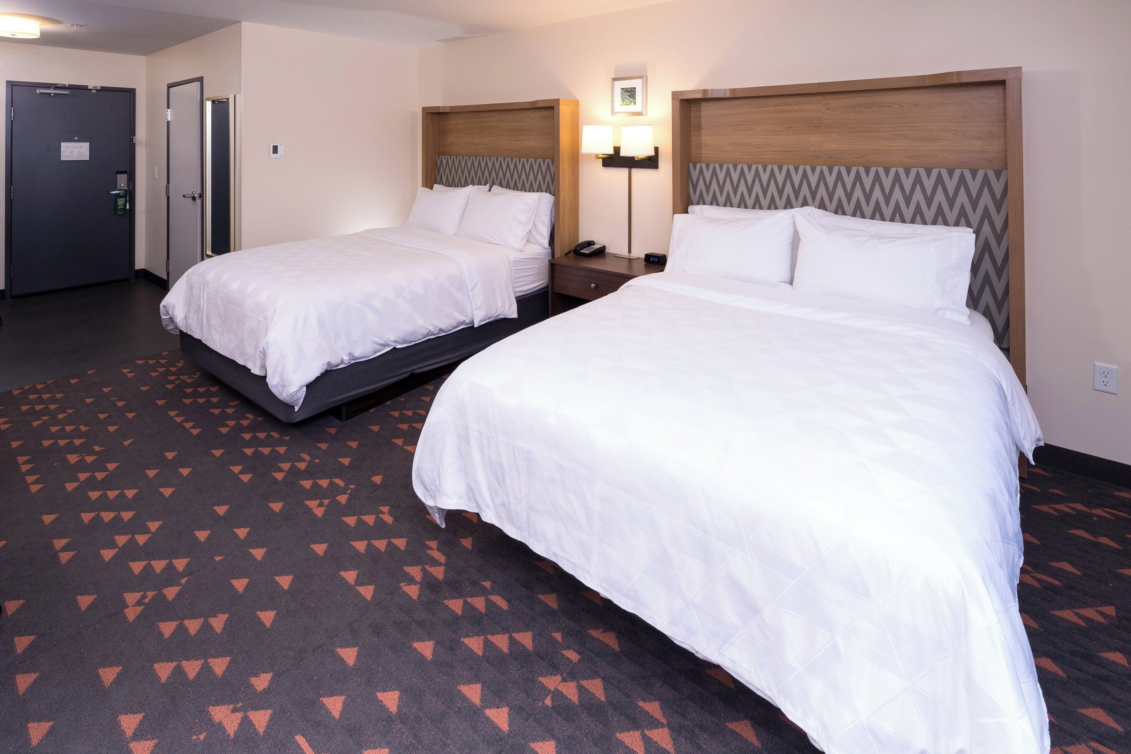 A hotel room with two beds, a nightstand, a wall-mounted lamp, and a patterned carpet.