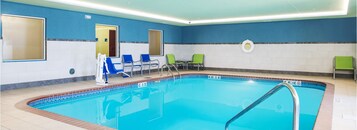 Una piscina interior con paredes de azulejos azules y blancos, una escalera de piscina y varias sillas alrededor del área.