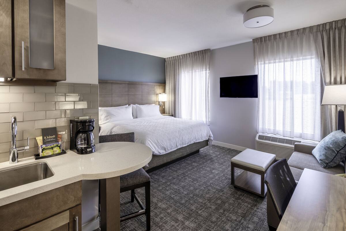 A hotel room with a bed, a desk, a chair, a coffee maker, a TV, and a window with curtains.