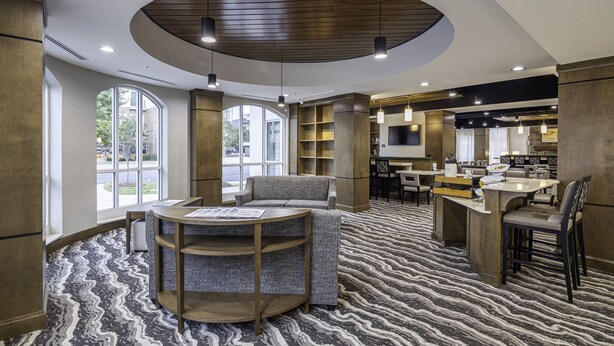 A modern hotel lobby with a circular wooden ceiling, large windows, a seating area with a round table, and a kitchen area in the background. - Staybridge Suites Montgomery - Downtown by IHG (Montgomery)