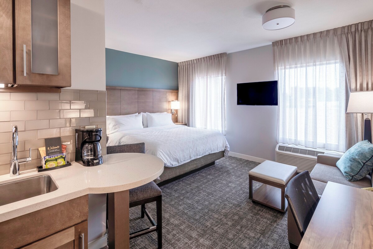 Habitación de hotel con cama, escritorio, silla, cafetera, televisor y ventana con cortinas.