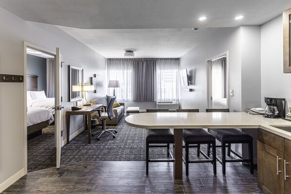 A modern hotel room with a bed, desk, chair, and kitchenette. - Staybridge Suites Montgomery - Downtown by IHG (Montgomery)