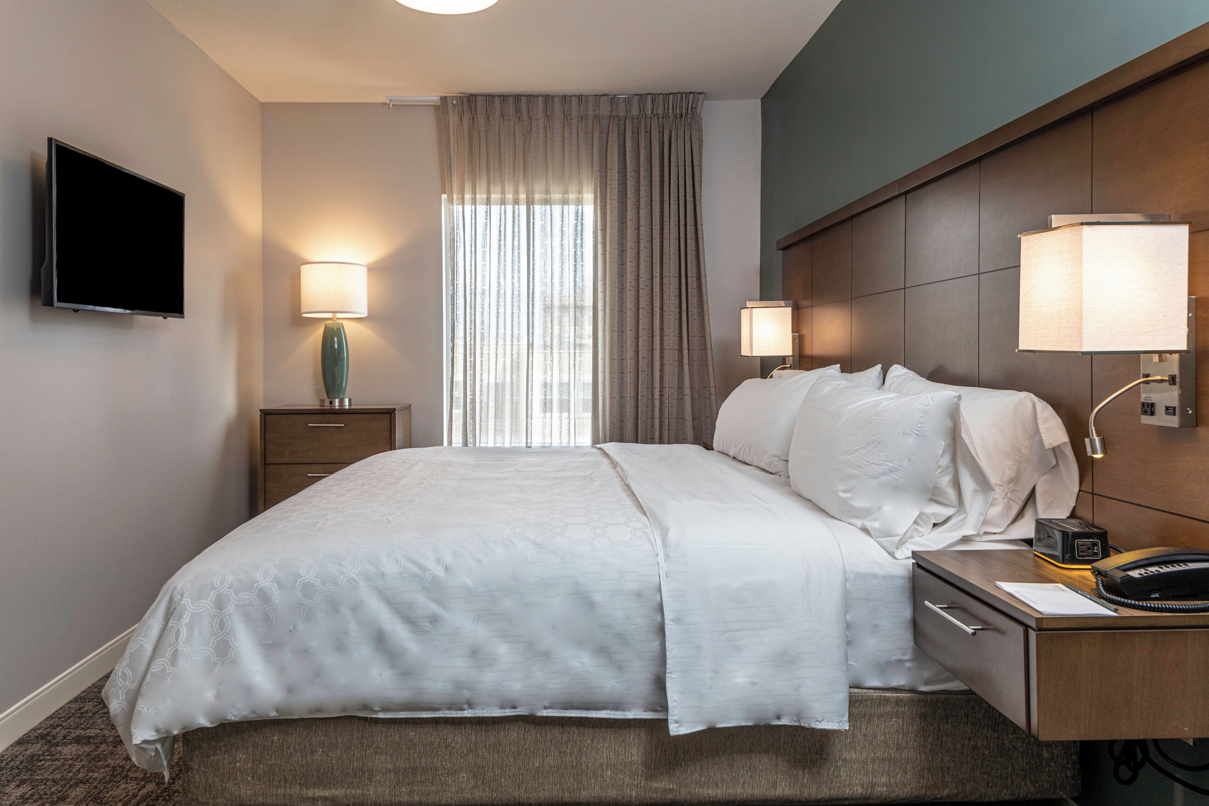 Habitación de hotel con cama, lámparas de noche, televisor de pantalla plana, escritorio con teléfono y ventana con cortinas.
