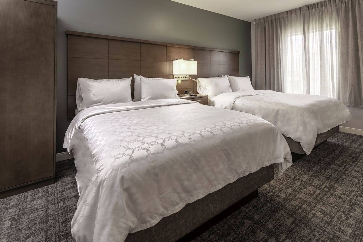 A hotel room with two beds, a headboard, a nightstand with a lamp, and a large window with curtains.