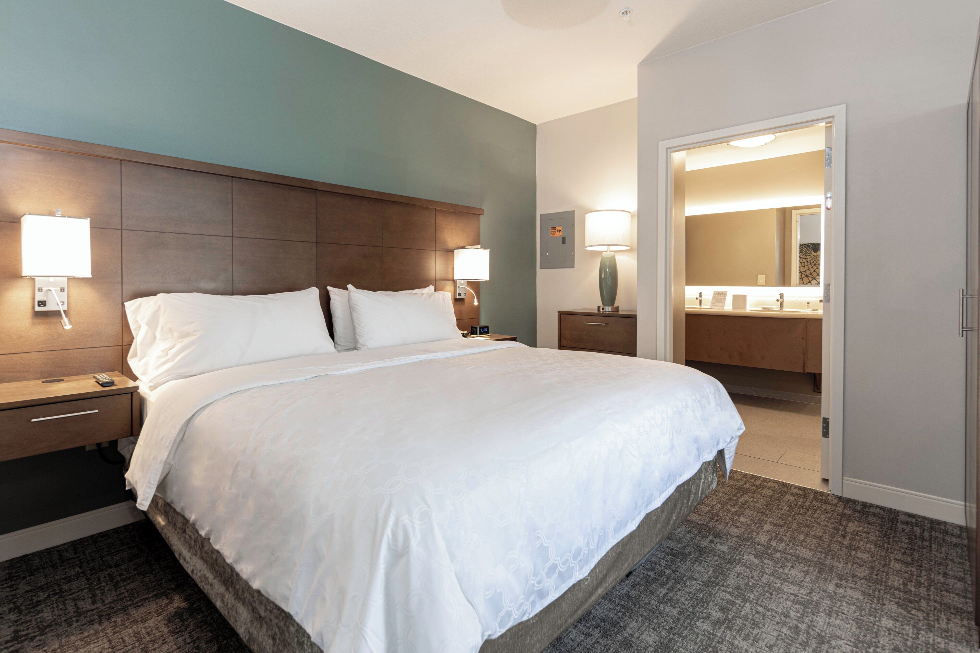 A hotel room with a large bed, bedside lamps, a nightstand, a dresser, and a bathroom visible through an open door.