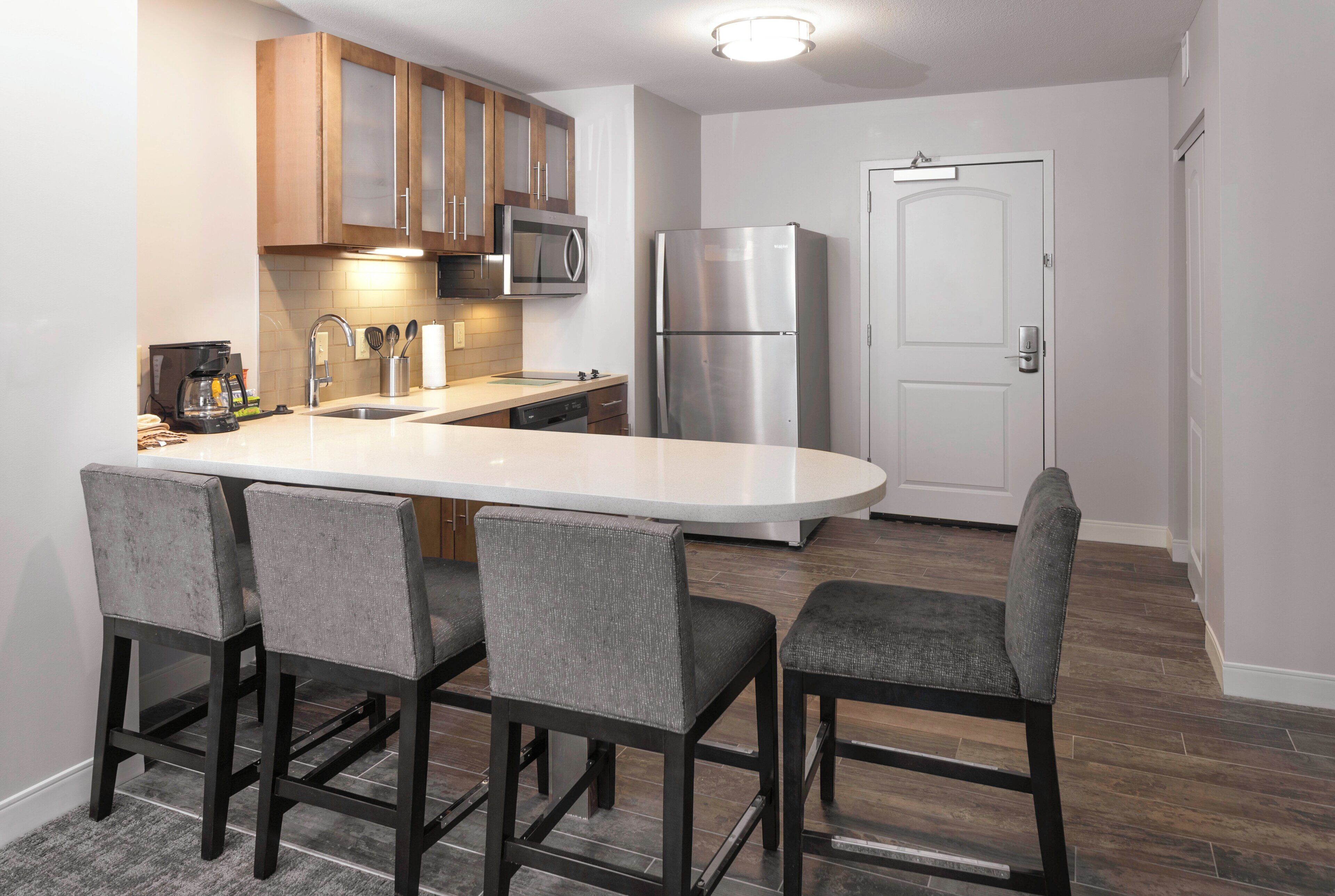 A modern kitchen with a central island, bar stools, stainless steel appliances, and a door in the background.