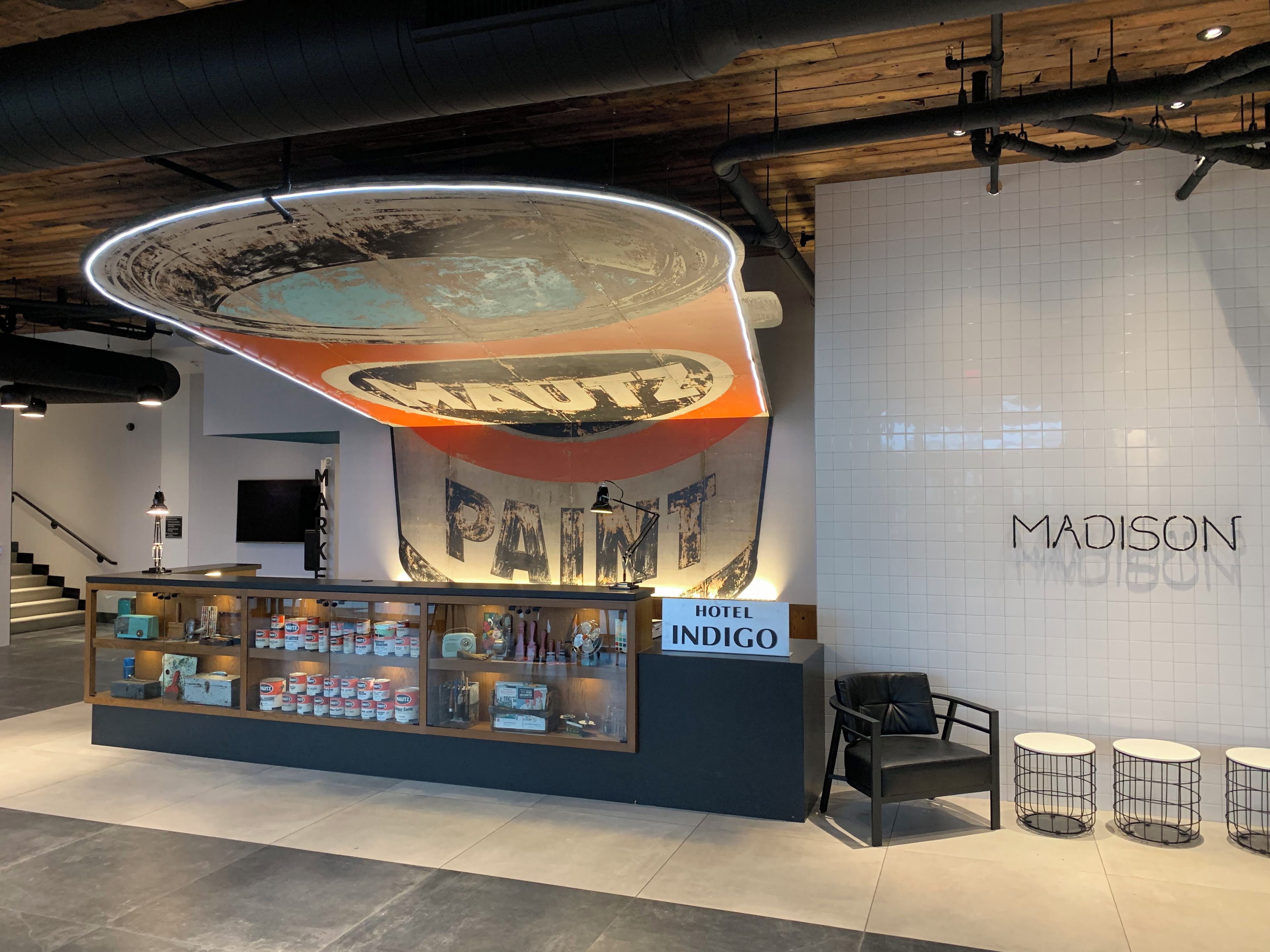 A hotel reception area with a large sign reading 'INDIGO' and a sign for 'HOTEL INDIGO.' There are display cases with various items, a staircase in the background, and a wall with a grid pattern.