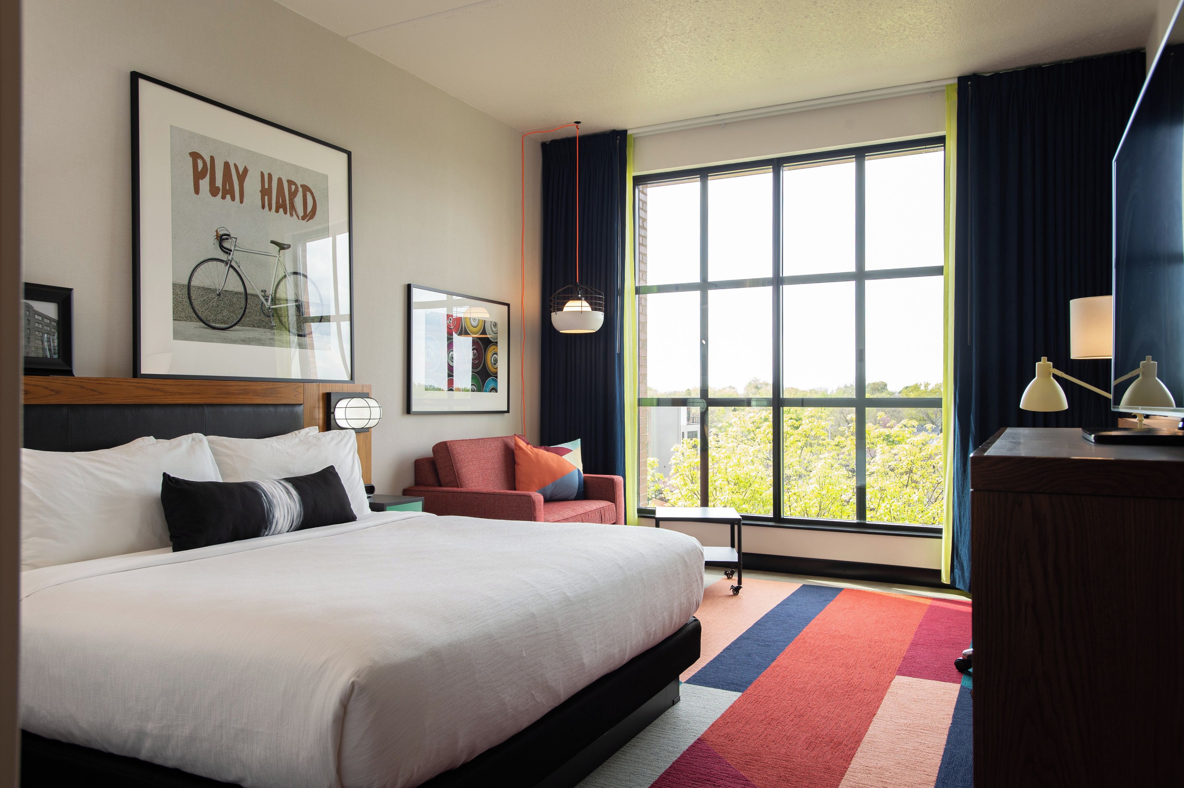 A hotel room with a large bed, a desk, a chair, a window with a view, and artwork on the walls.