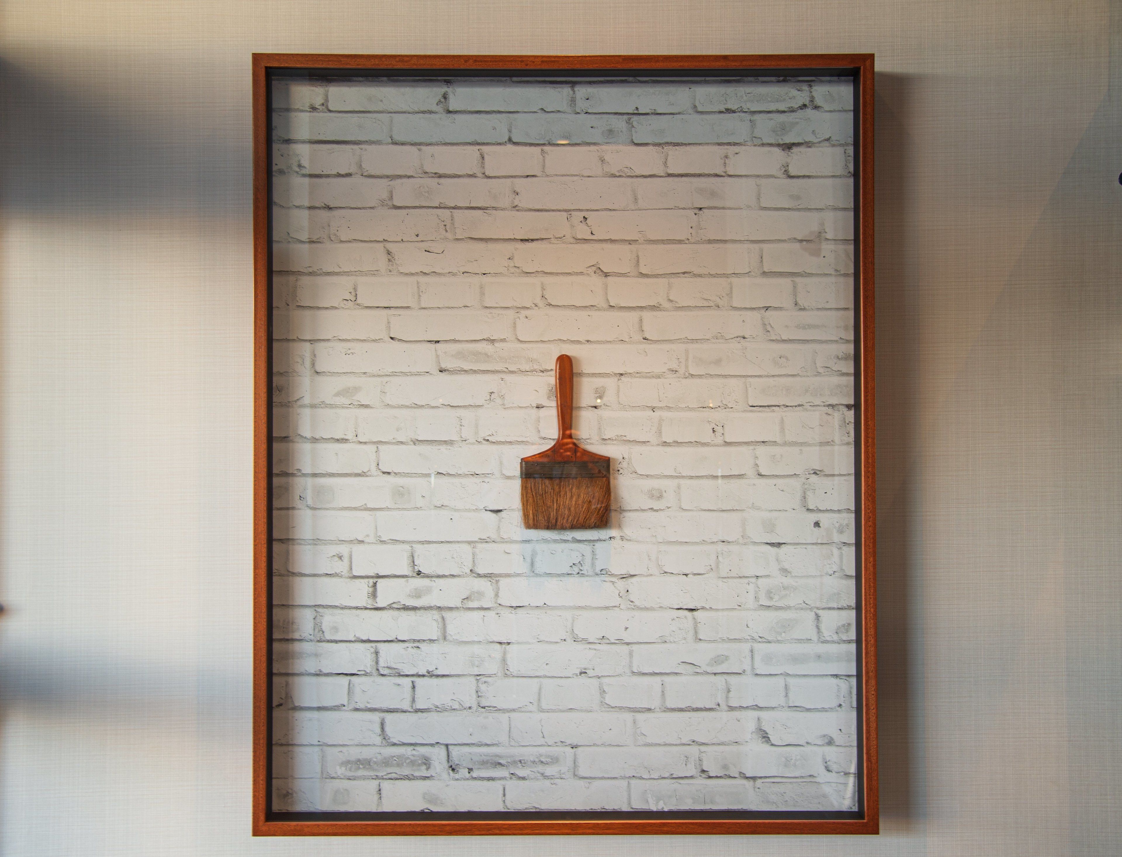 A framed painting of a wooden brush on a white brick wall.
