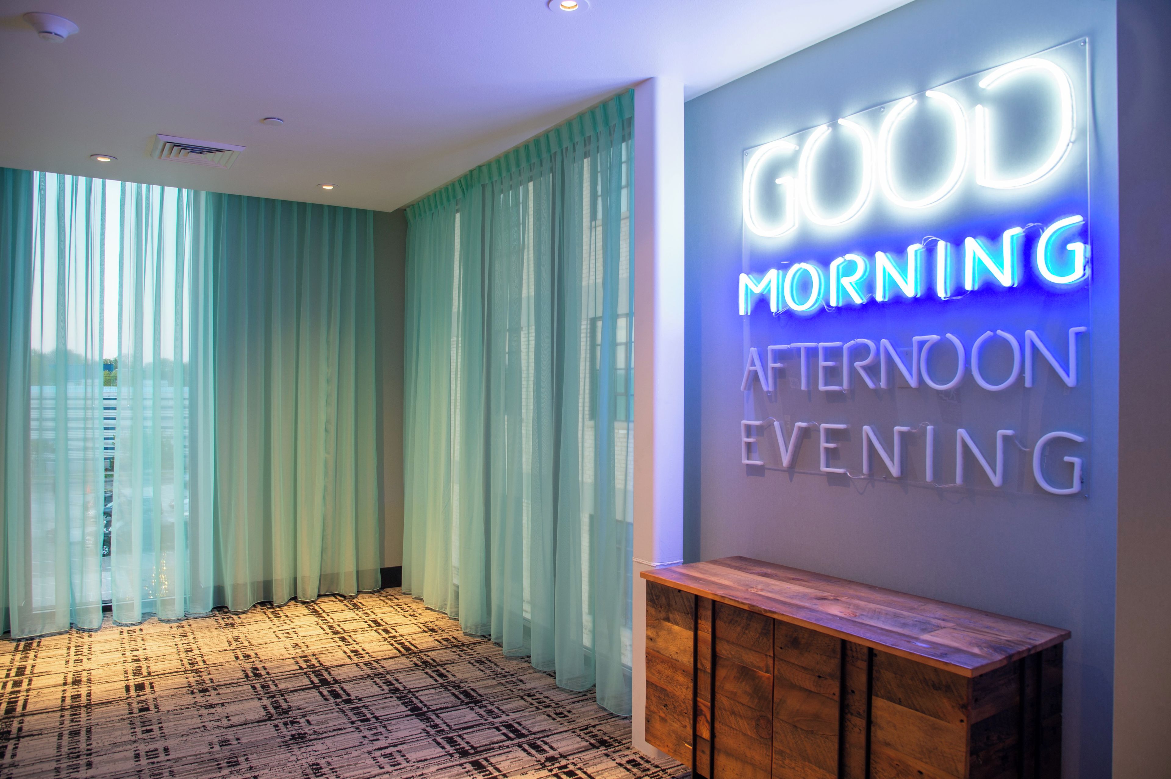A hotel lobby with a neon sign reading 'GOOD MORNING APOSTOL EVENING' and a patterned carpet.