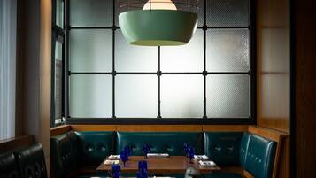 Um restaurante com disposição em cabines, uma cabine estofada em verde, uma mesa de madeira com copos de vidro azul e um lustre pendente.