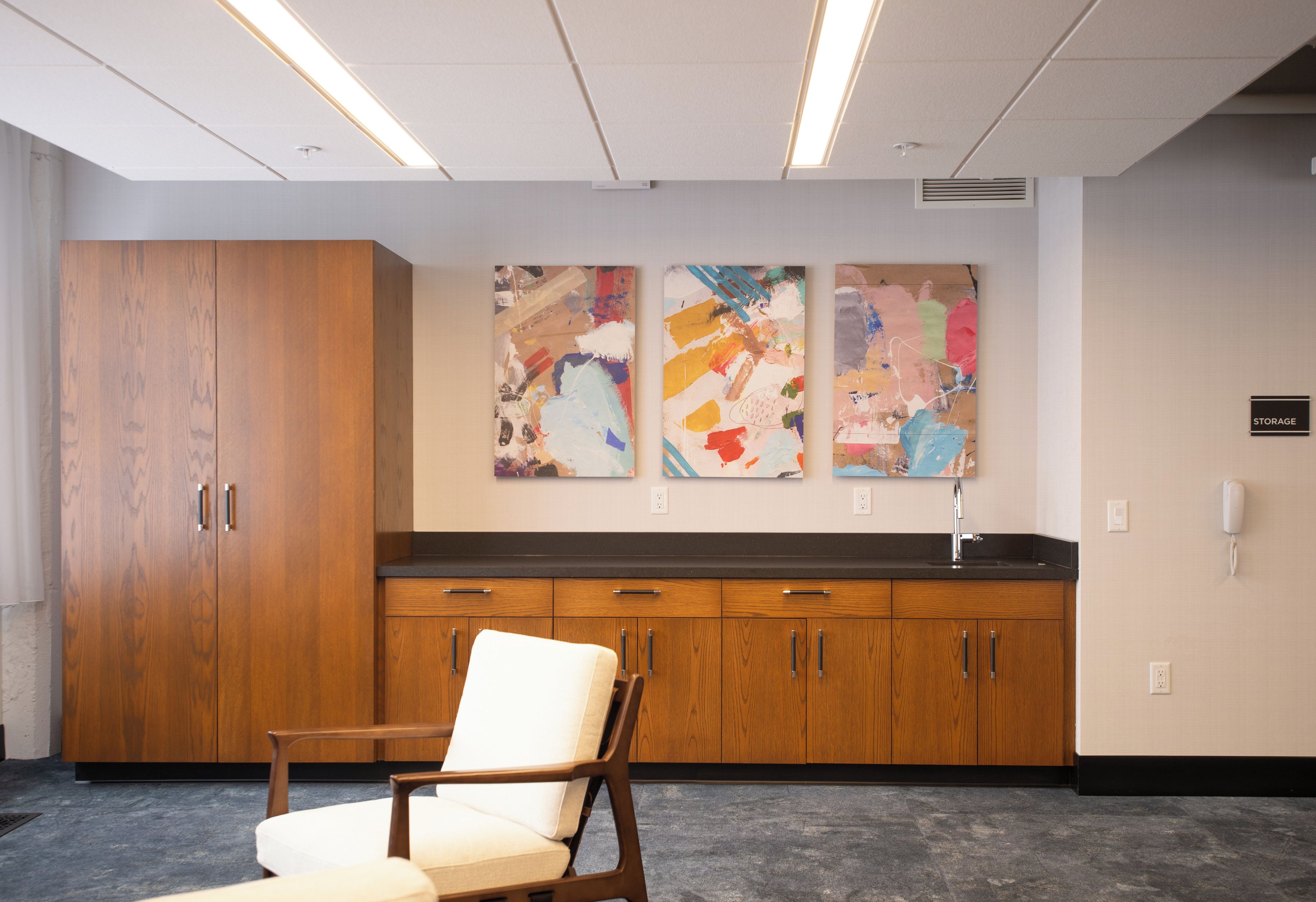 A waiting area with a wooden cabinet, a white cushioned chair, and abstract paintings on the wall.