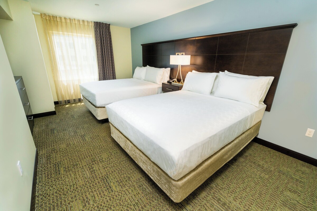 A hotel room with two beds, a nightstand with a lamp, a window with curtains, and a carpeted floor.