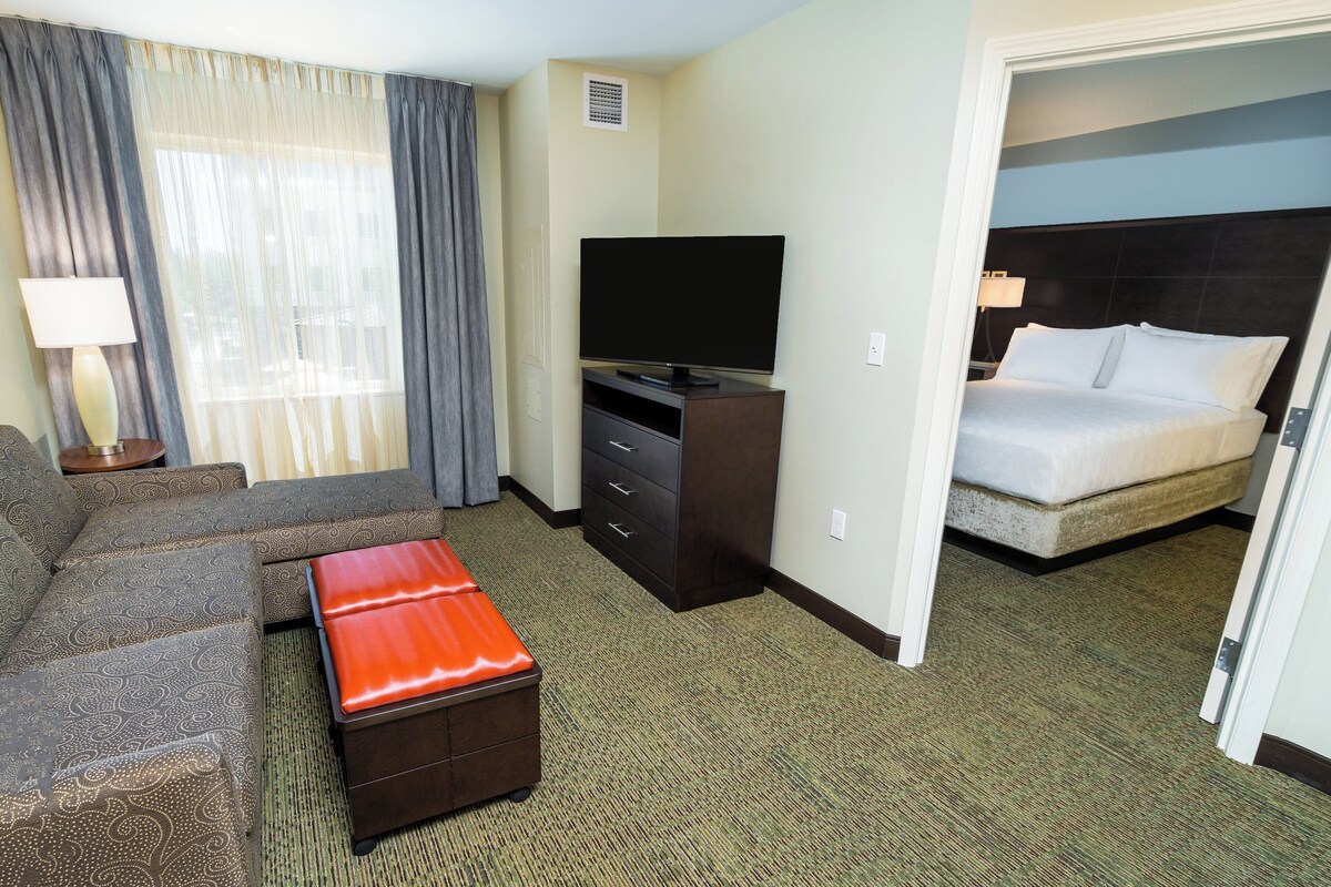 A hotel room with a sofa, a television on a stand, a bed, and a red ottoman.
