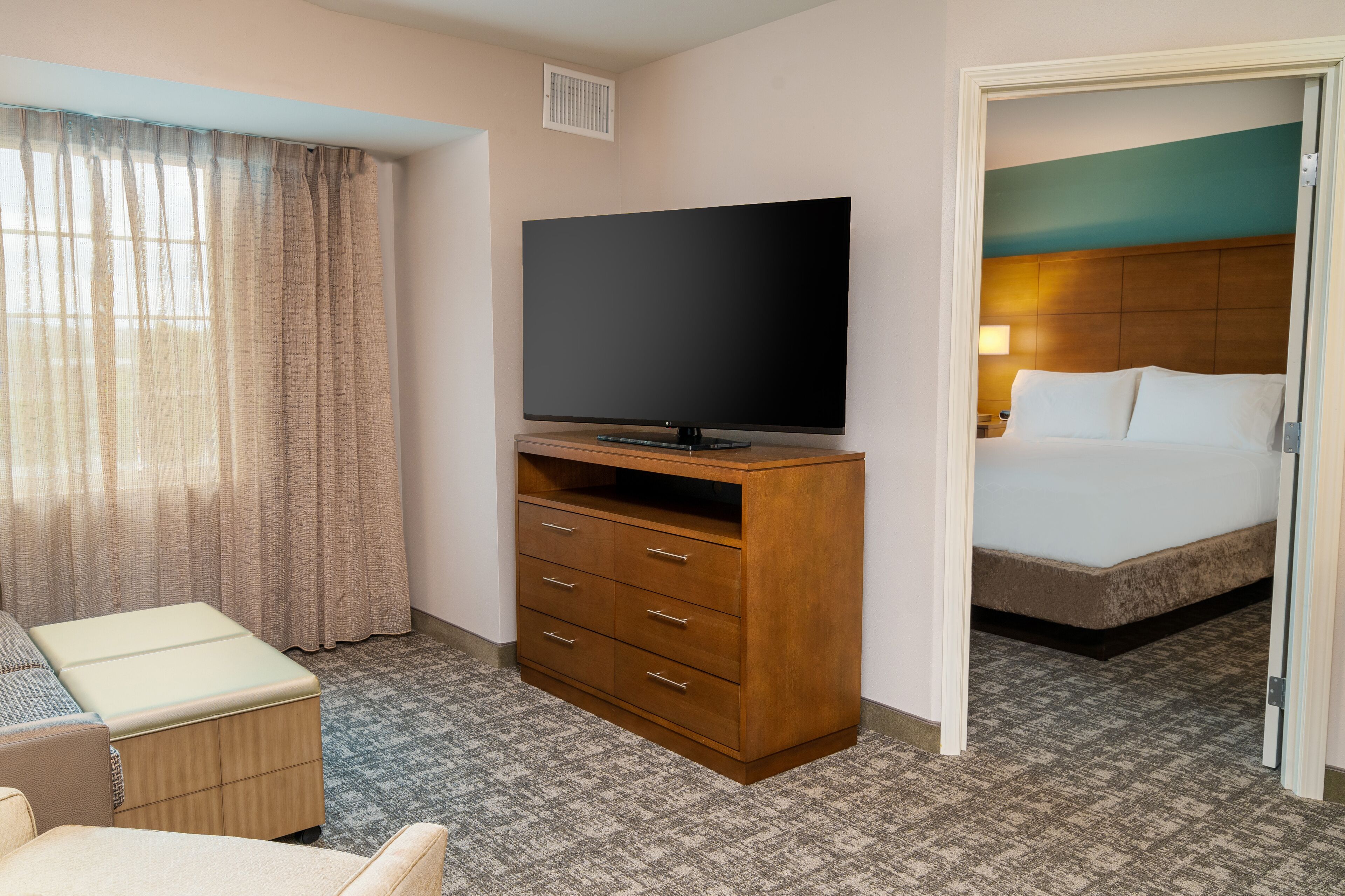 A hotel room with a bed, a television on a wooden stand, a desk with a chair, and a window with curtains.
