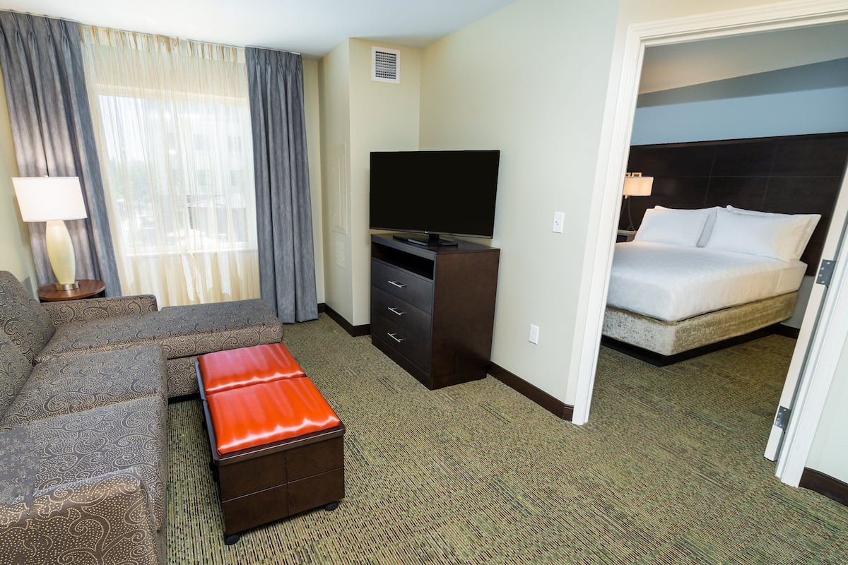 A hotel room with a sofa, a television on a stand, a bed, and a red ottoman.