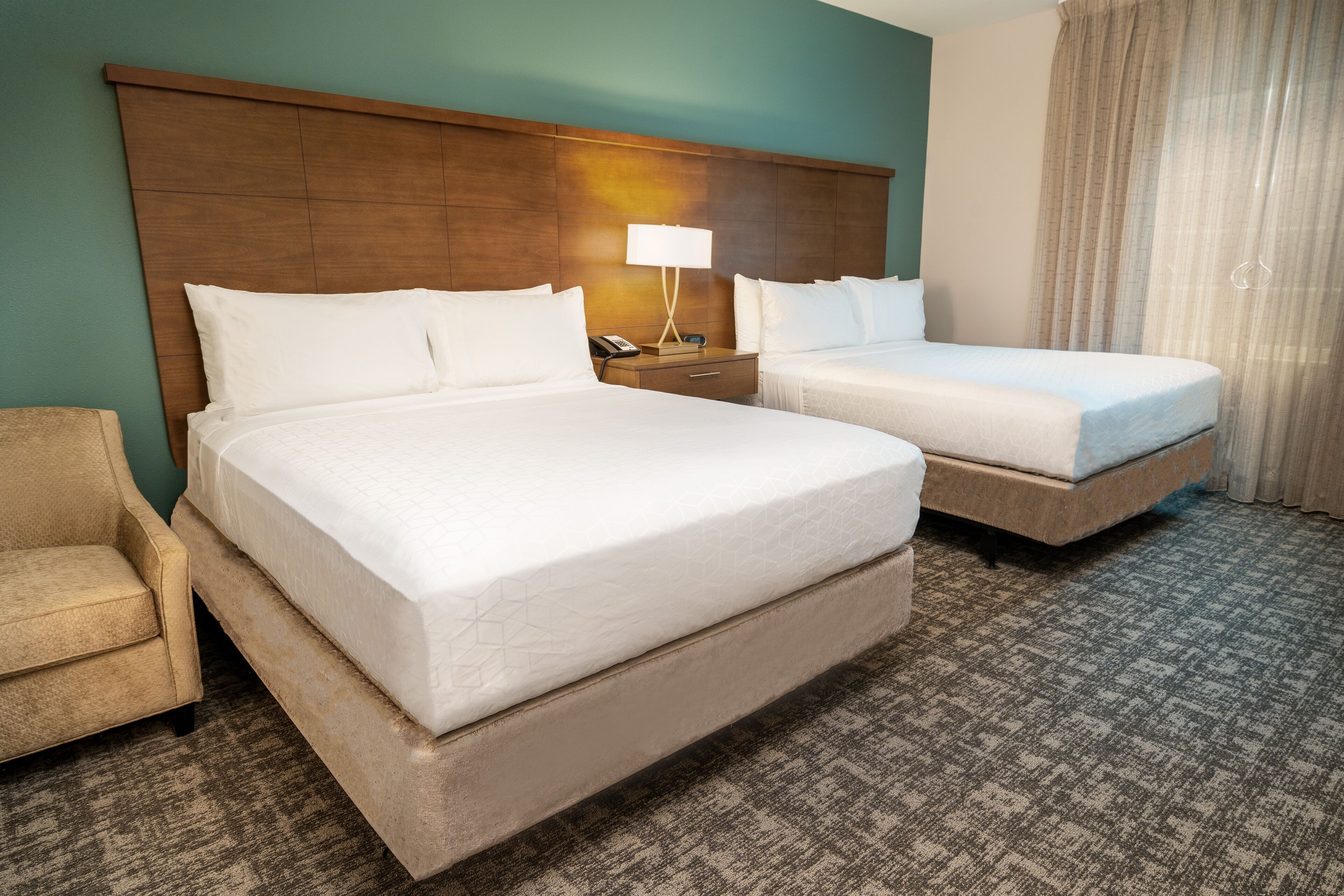 A hotel room with two beds, a nightstand with a lamp, a chair, and a patterned carpet.