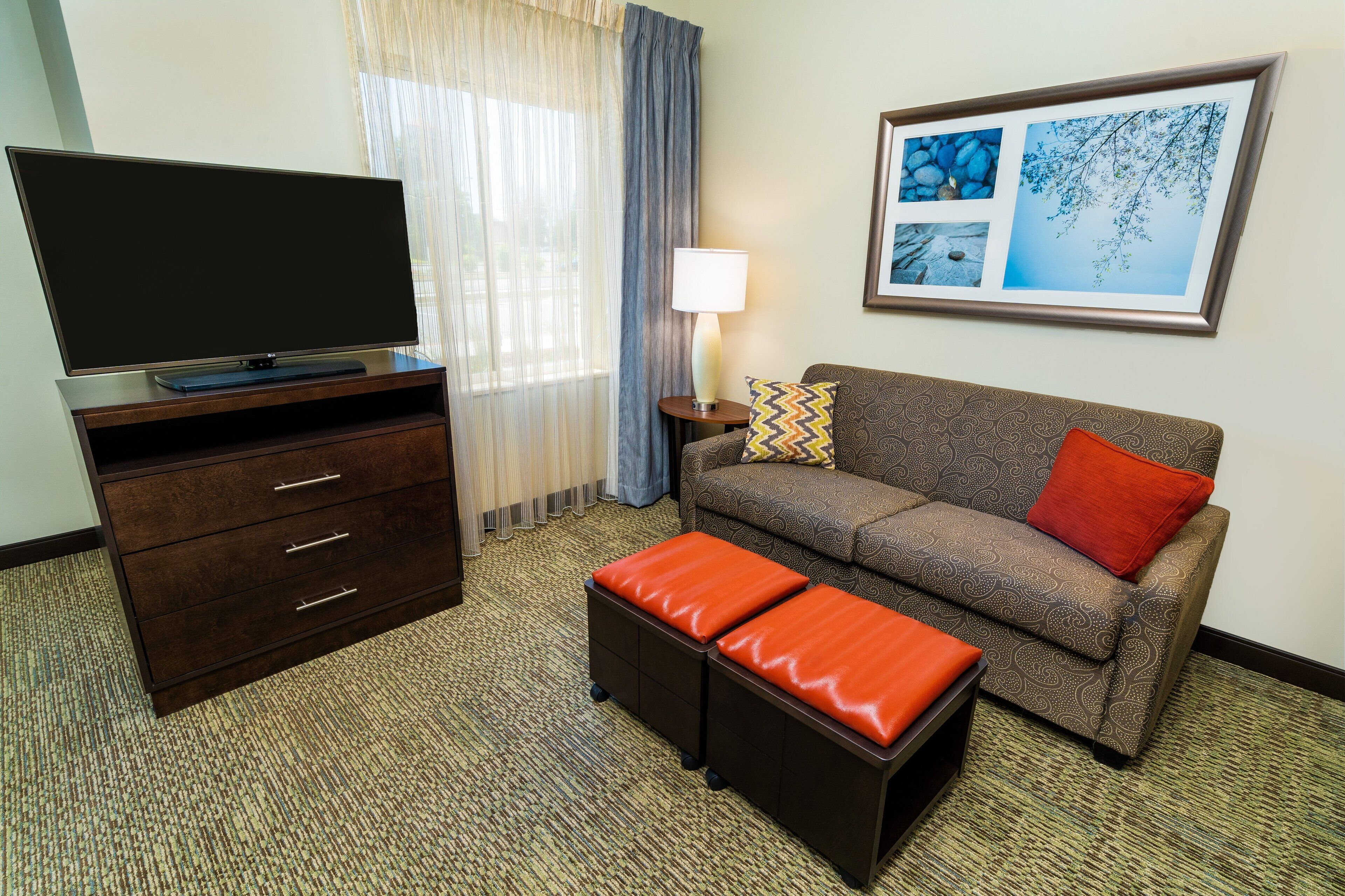 Ein Hotelzimmer mit Flachbildfernseher auf einem Holzkabinett, einer Couch mit gemusterten Kissen, einem kleinen Tisch mit Lampe und einem gerahmten Bild an der Wand.