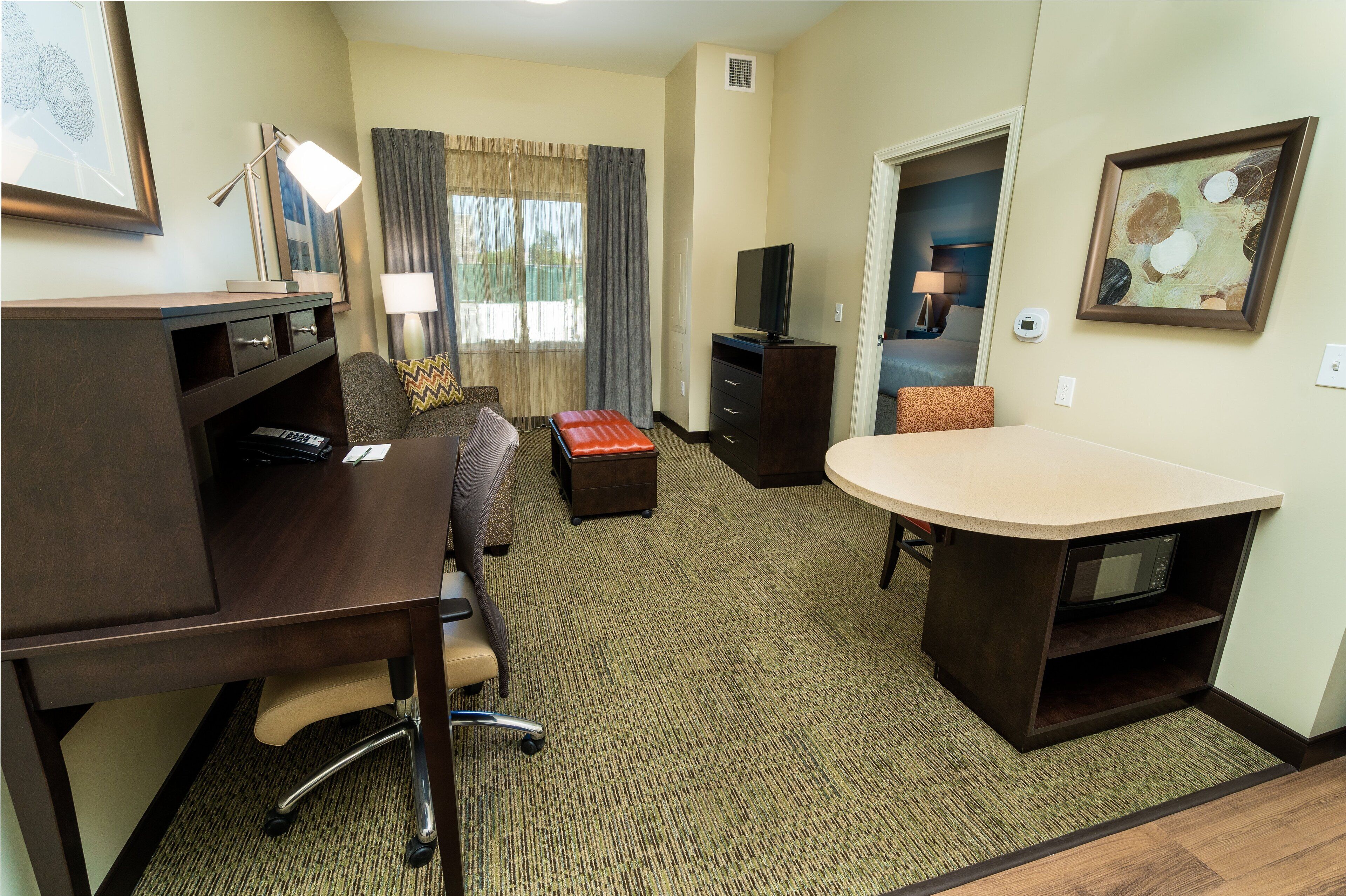 A hotel room with a desk, a sofa, a television, a microwave, and a painting on the wall.