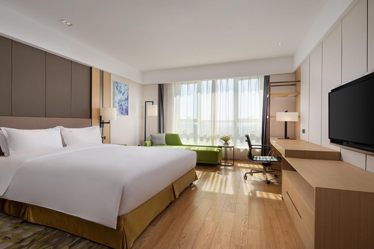 A hotel room with a large bed, a desk, a chair, a television, and a window with curtains.