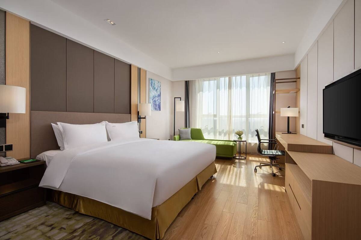 Een moderne hotelkamer met een groot bed, een bureau met een flatscreen televisie, een groene fauteuil en een bijzettafeltje met een lamp.