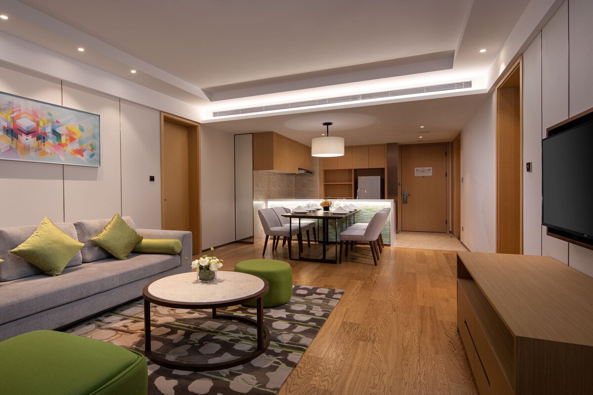 A modern living room with a sofa, a coffee table, and a TV. There is a dining area with chairs and a table. The room has wooden flooring and a large painting on the wall.