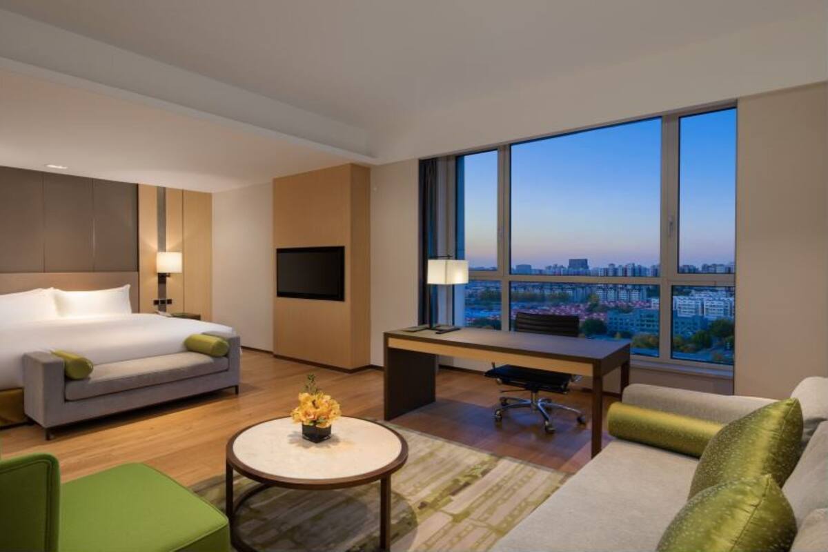 Een moderne hotelkamer met een bed, een bureau, een groene stoel, een bank en een groot raam met uitzicht op de stad.