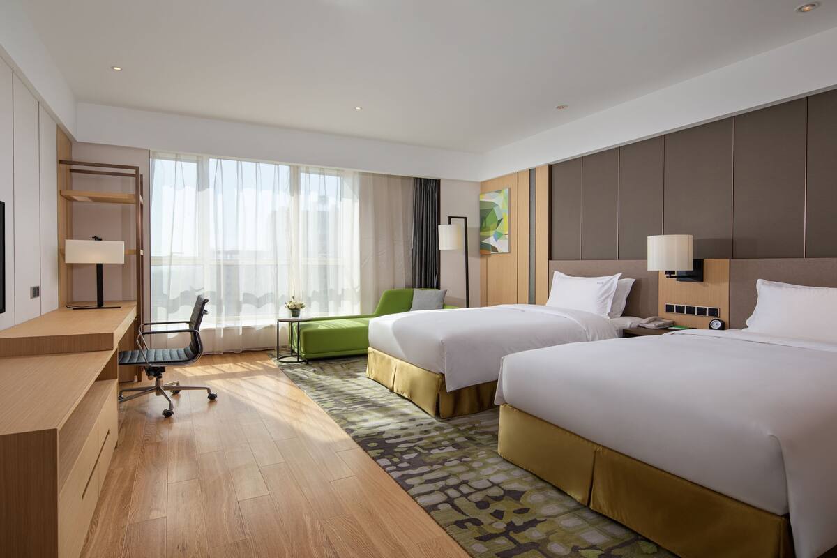 Een hotelkamer met twee bedden, een bureau met een televisie, een stoel, een lamp en een groot raam.