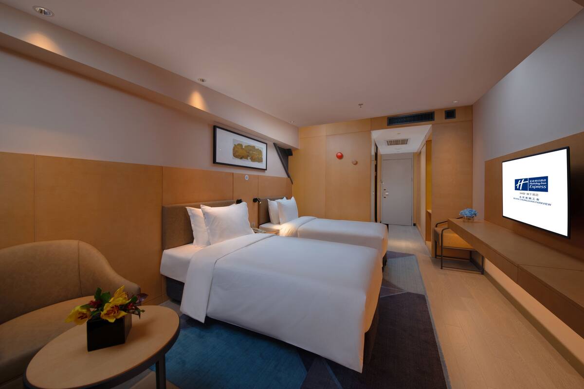 A hotel room with two beds, a desk, a chair, a TV, and a small table with flowers.