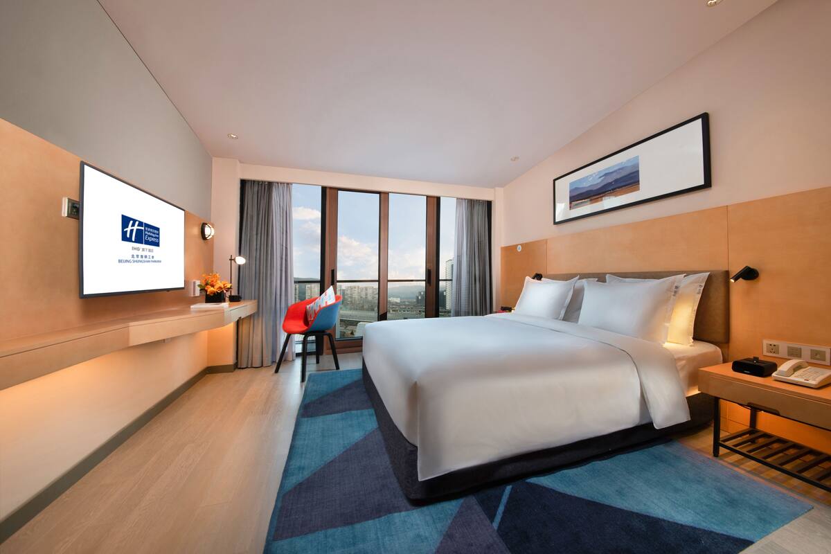 A modern hotel room with a large bed, a desk with a chair, a TV mounted on the wall, and a large window offering a city view.