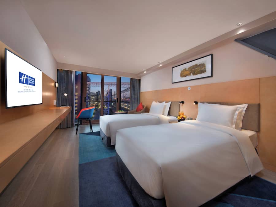 Habitación de hotel con dos camas, un escritorio, una silla y un amplio ventanal con vistas a la ciudad.