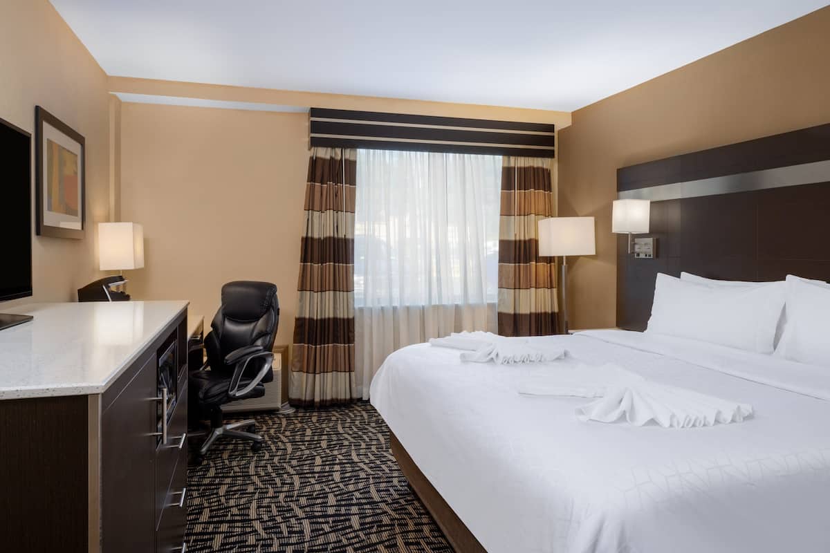 A hotel room with a bed, two bedside lamps, a desk with a chair, a TV, and a patterned carpet.