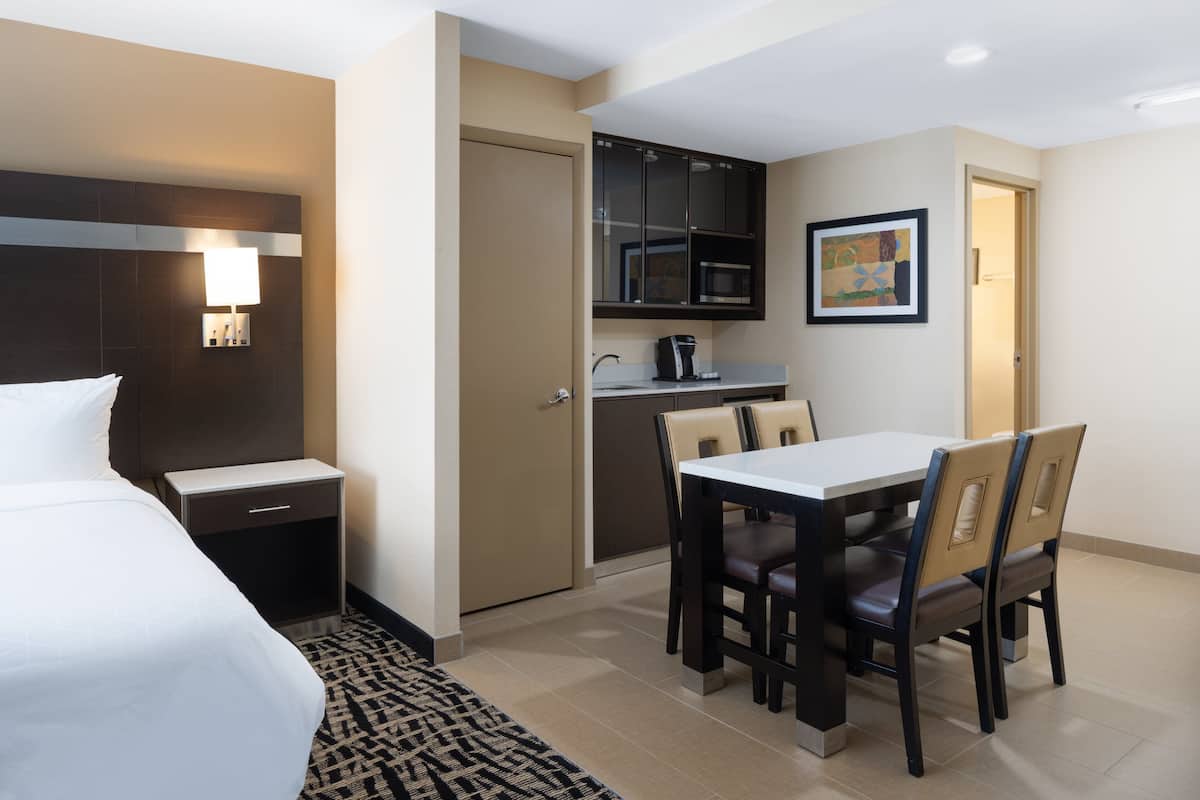 A hotel room with a bed, a desk, a chair, a small kitchenette, and a painting on the wall.