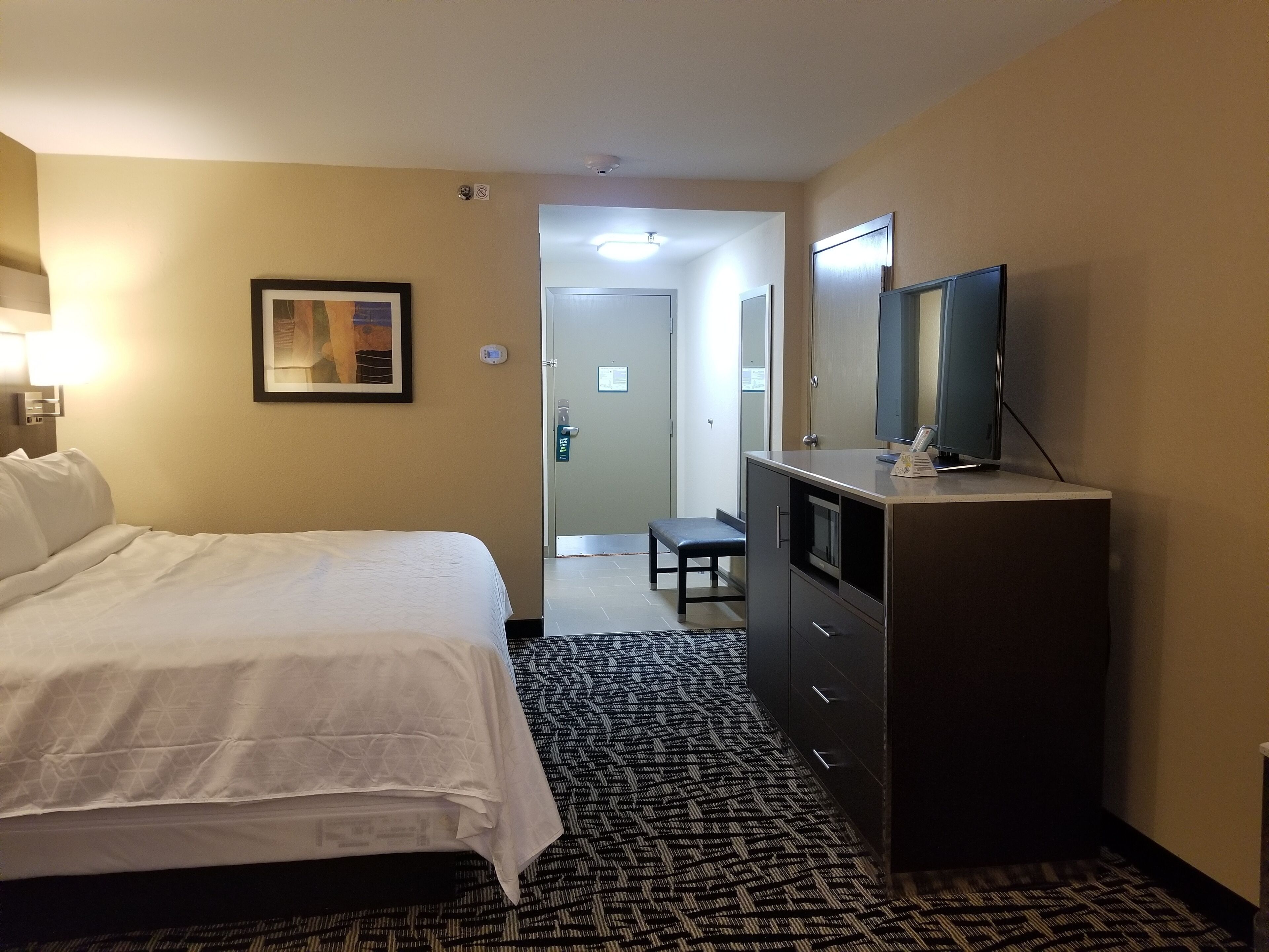Habitación de hotel con cama, escritorio con televisor, cuadro en la pared y puerta que da a otra habitación.