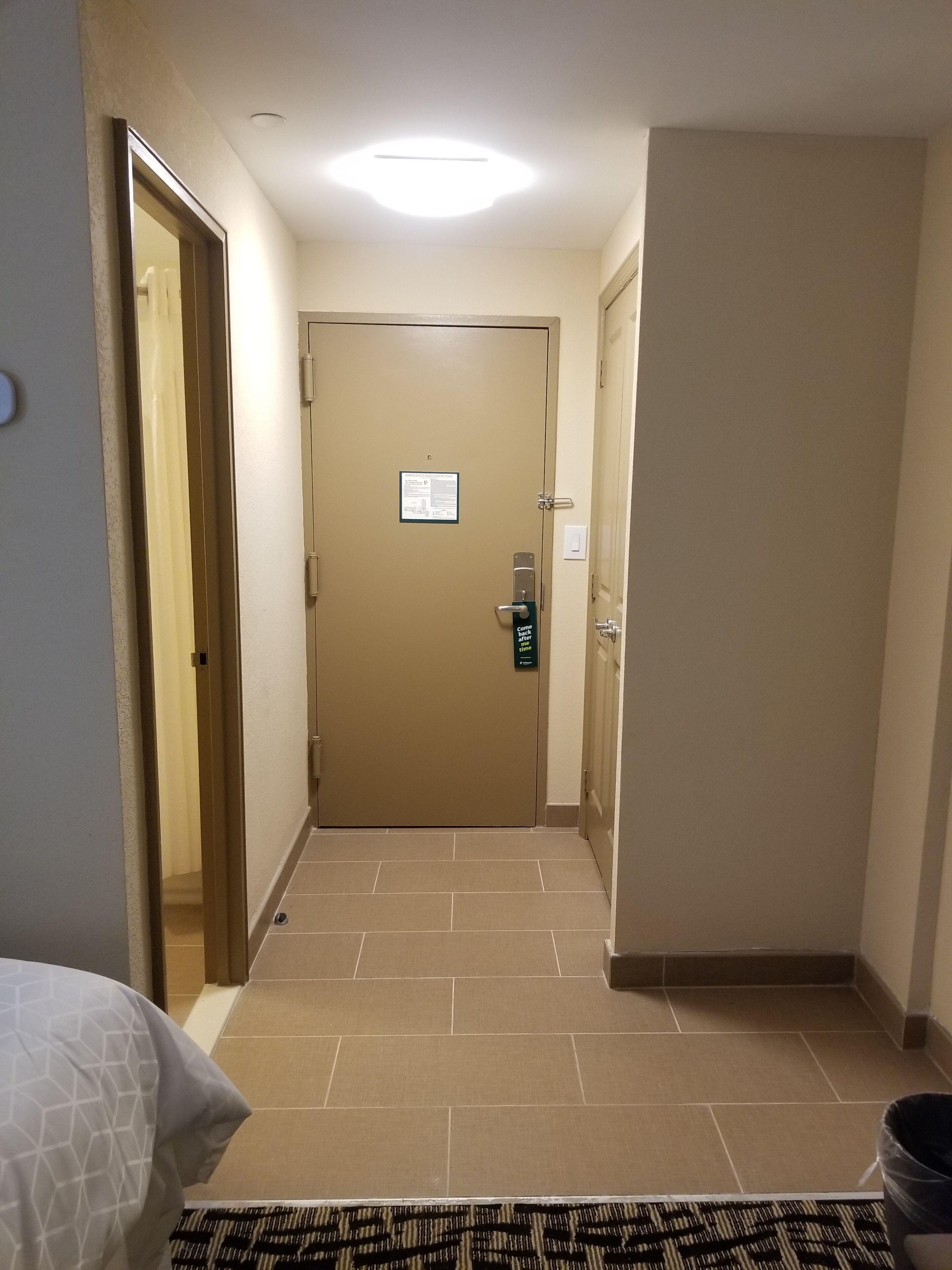 A hotel hallway with a closed door, a bed with a geometric-patterned cover, and a trash bin.