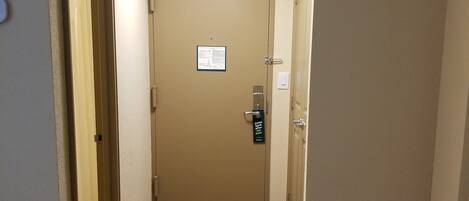 A hotel hallway with a closed door, a bed with a geometric-patterned cover, and a trash bin.