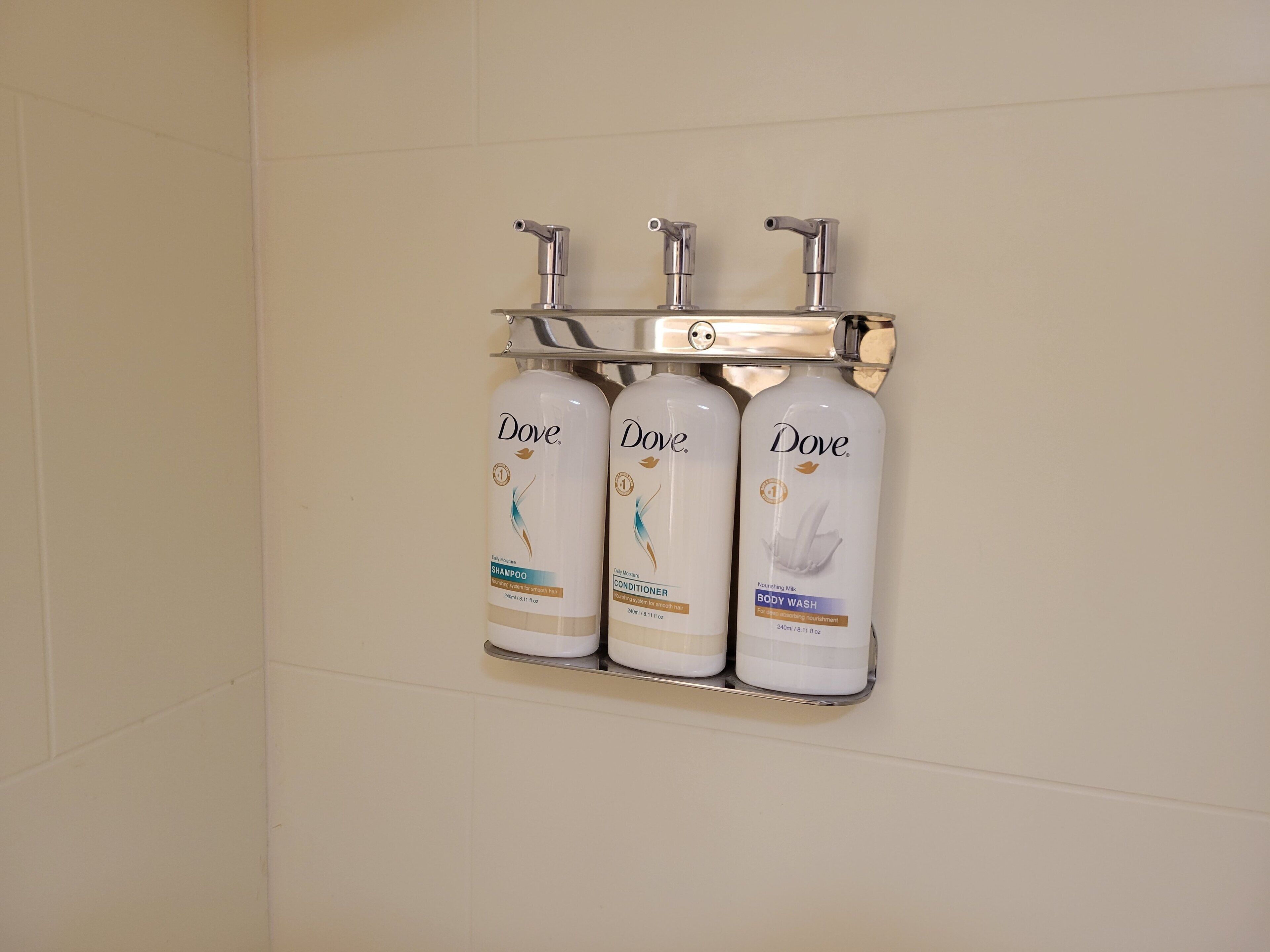 Casa de banho com três produtos Dove: champô, condicionador e gel de banho, montados num dispensador fixado na parede.