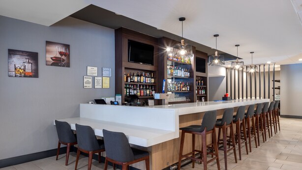 A modern bar area with a long counter, multiple bar stools, and a variety of liquor bottles. - Holiday Inn Poughkeepsie by IHG (Poughkeepsie)