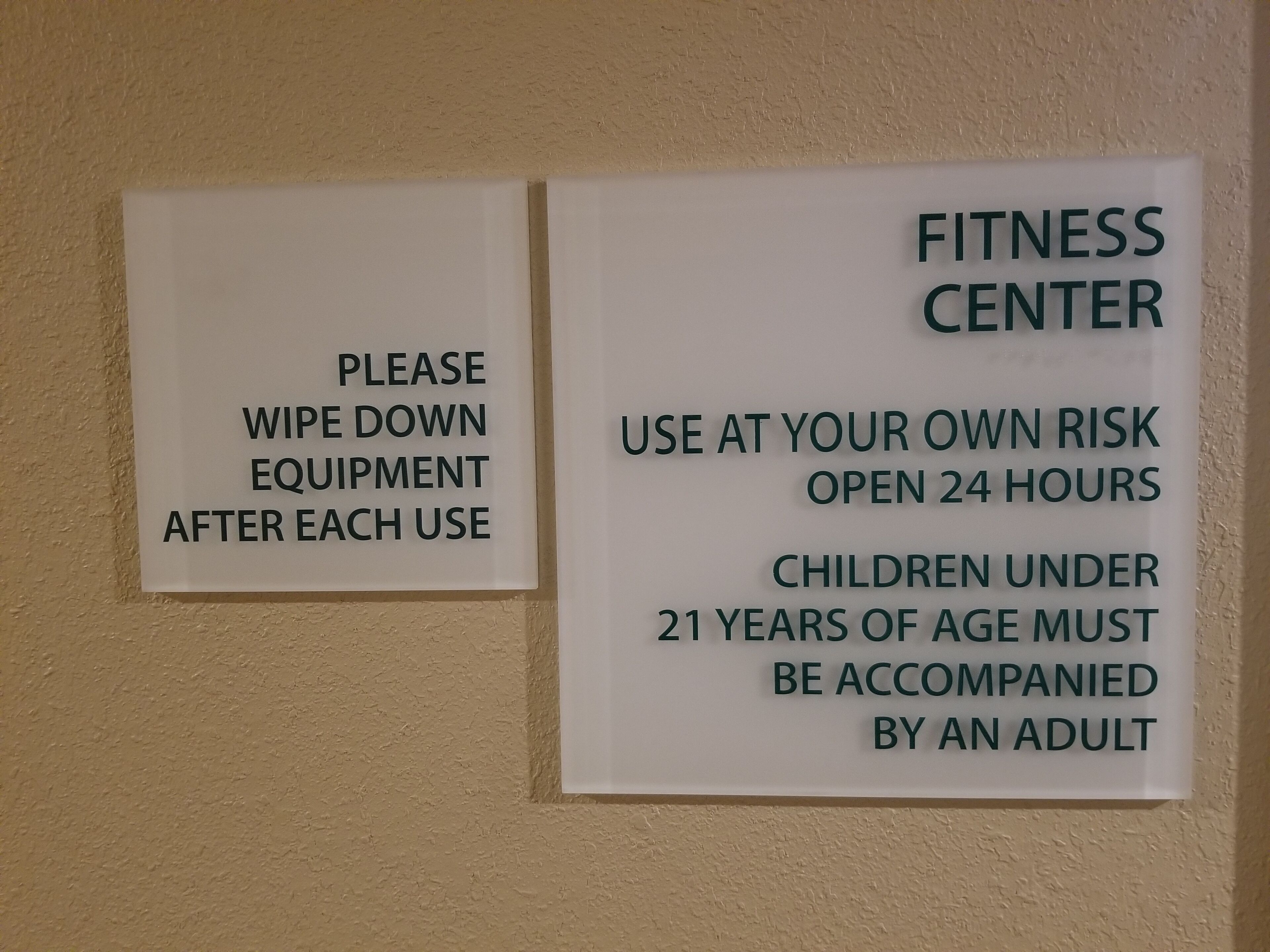 Two signs on a wall. One sign says "Please wipe down equipment after each use." The other sign says "Fitness Center use at your own risk open 24 hours children under 21 years of age must be accompanied by an adult."