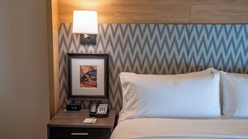 Habitación de hotel con cama, mesita de noche con lámpara, un cuadro enmarcado, un reloj digital y un teléfono.