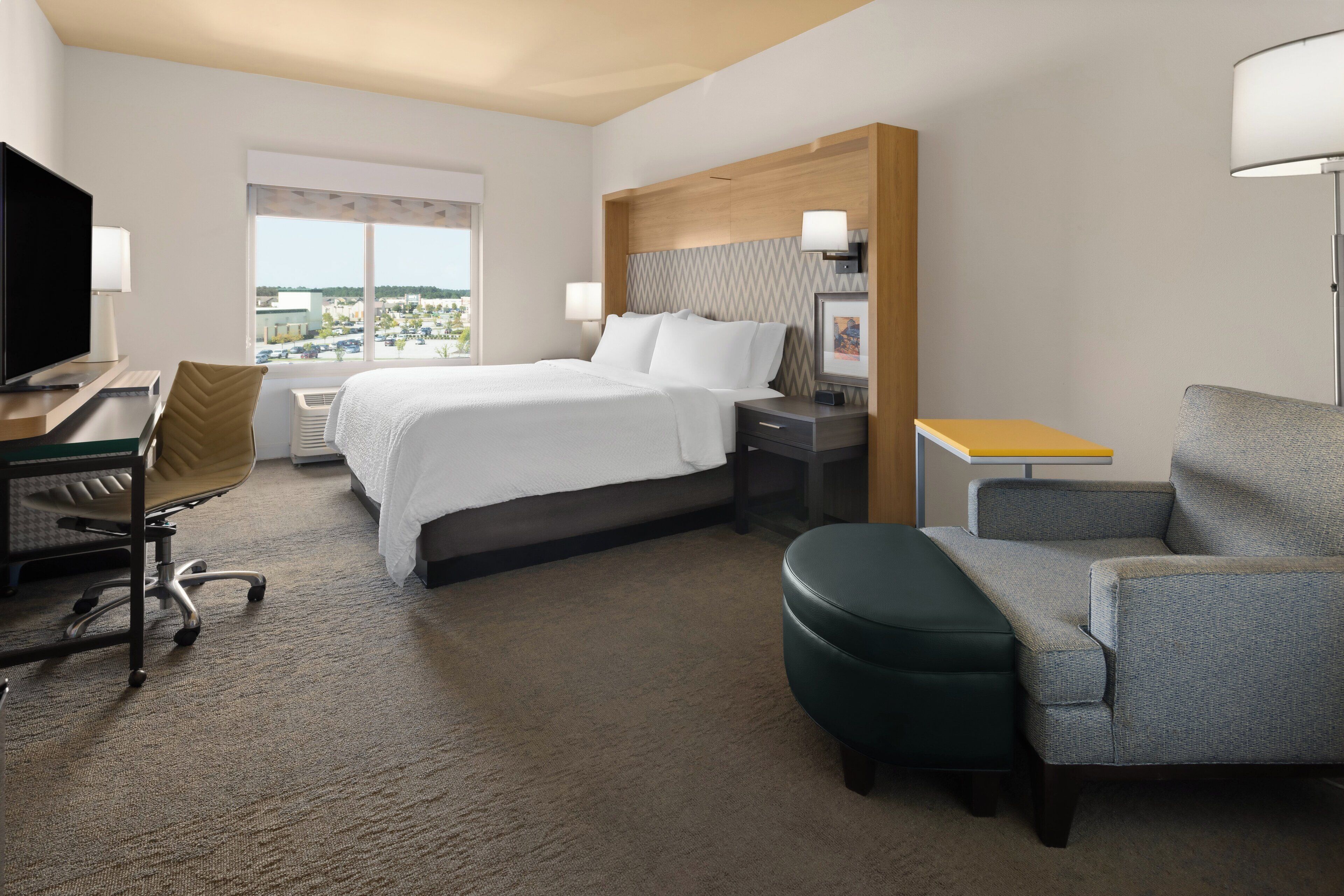 A hotel room with a bed, a desk with a chair, a TV, a lamp, and a view of the city.