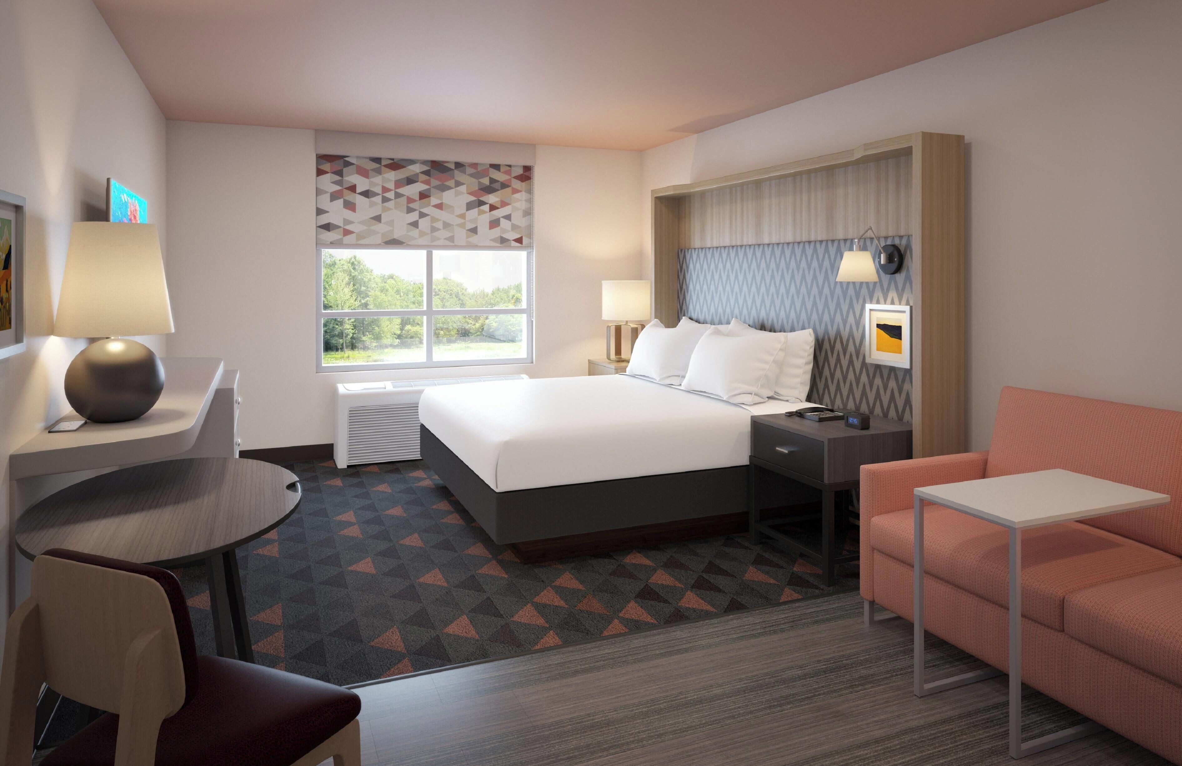 A hotel room with a bed, a desk, a chair, a sofa, and a window with a view of trees.